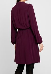 Långärmad, lila klänning med en rynkad halsringning och elastisk midja. Tillverkad av mjukt tyg, med en flödande silhuett och en knälång nederdel.