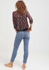 Blouse noire à motifs floraux avec manches trois-quarts, associée à un jean skinny bleu clair, mettant en valeur un design fendu au dos et des chaussures plates de couleur neutre.