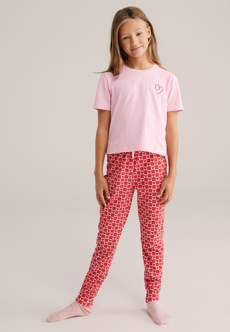 T-shirt in cotone rosa con ricamo a forma di cuore, abbinato a pantaloni da pigiama rossi con una fantasia a cuori bianchi. Il modello indossa calzini rosa chiaro.