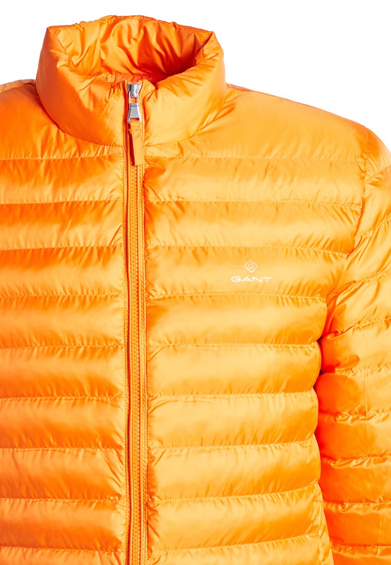 Chaqueta acolchada naranja con cuello alto, diseño de acolchado horizontal, cremallera frontal y logotipo en el lado izquierdo. Textura lisa y brillante.