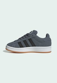 Zapatilla de ante gris con tres franjas negras, media suela blanca y suela de goma marrón. Punta redondeada y cordones planos. Diseño atlético clásico.