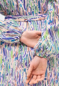 Mains croisées dans le dos, portant une robe à manches longues avec des poignets élastiques et un motif abstrait multicolore en bleu, vert, beige et violet.