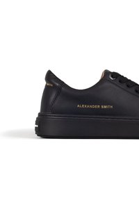 Sneaker in pelle nera con suola piatta, scritta dorata sul lato che legge "Alexander Smith" e lacci neri. Texture liscia e design minimalista.