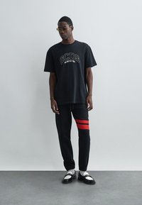 Tricou GCDS negru cu logo, pantaloni negri cu dungi roșii și loafers negri cu alb. Accesoriile includ ochelari de soare galbeni.