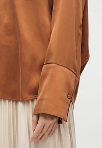 Κοντινό πλάνο ενός ατόμου που φορά καφέ πουκάμισο με μανσέτα με κουμπί και κρεμ πλισέ φούστα.