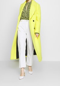 Dlouhý jasně žlutý vlněný kabát, černá květinová blůza, bílé zkrácené džíny a bílé lodičky na špičce. Hladká textura, dvouřadové zapínání.