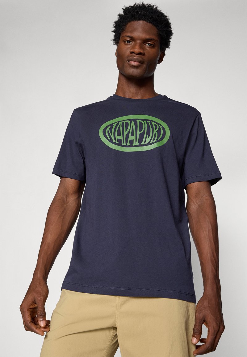 T-shirt bleu marine en coton avec un col rond, arborant un logo ovale vert "NAPAPIJRI" imprimé sur le devant. Manches courtes.