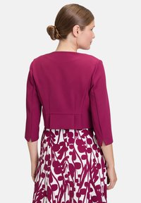 Dopasowana, skrócona kurtka w głębokim magencie z rękawami o długości trzech czwartych. Tył ma gładkie wykończenie oraz detale szwów. W zestawie ze wzorzystą spódnicą.