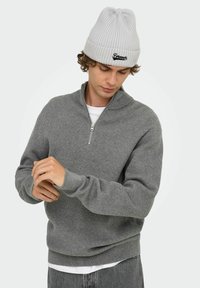 Grauer Strickpullover mit einem halben Reißverschluss am Kragen, gerippter Textur und langen Ärmeln. Kombiniert mit einer hellgrauen gerippten Mütze.