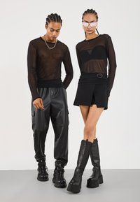 Schwarzes, transparentes Langarmoberteil und schwarze Cargo-Hosen an einem männlichen Model; schwarzes, eng anliegendes Kleid mit Ausschnitt-Detail und kniehohen Stiefeln an einem weiblichen Model.