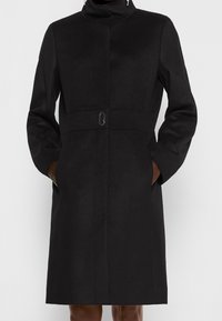 Cappotto di lana nera con colletto alto, maniche lunghe e chiusura con bottoni frontali. Presenta tasche laterali e un design aderente in vita. Tessuto liscio.