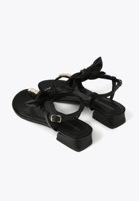 Black sandals with a satin finish, featuring a bow accent and a pearl-like detail on the strap. They have a low block heel and an adjustable buckle.