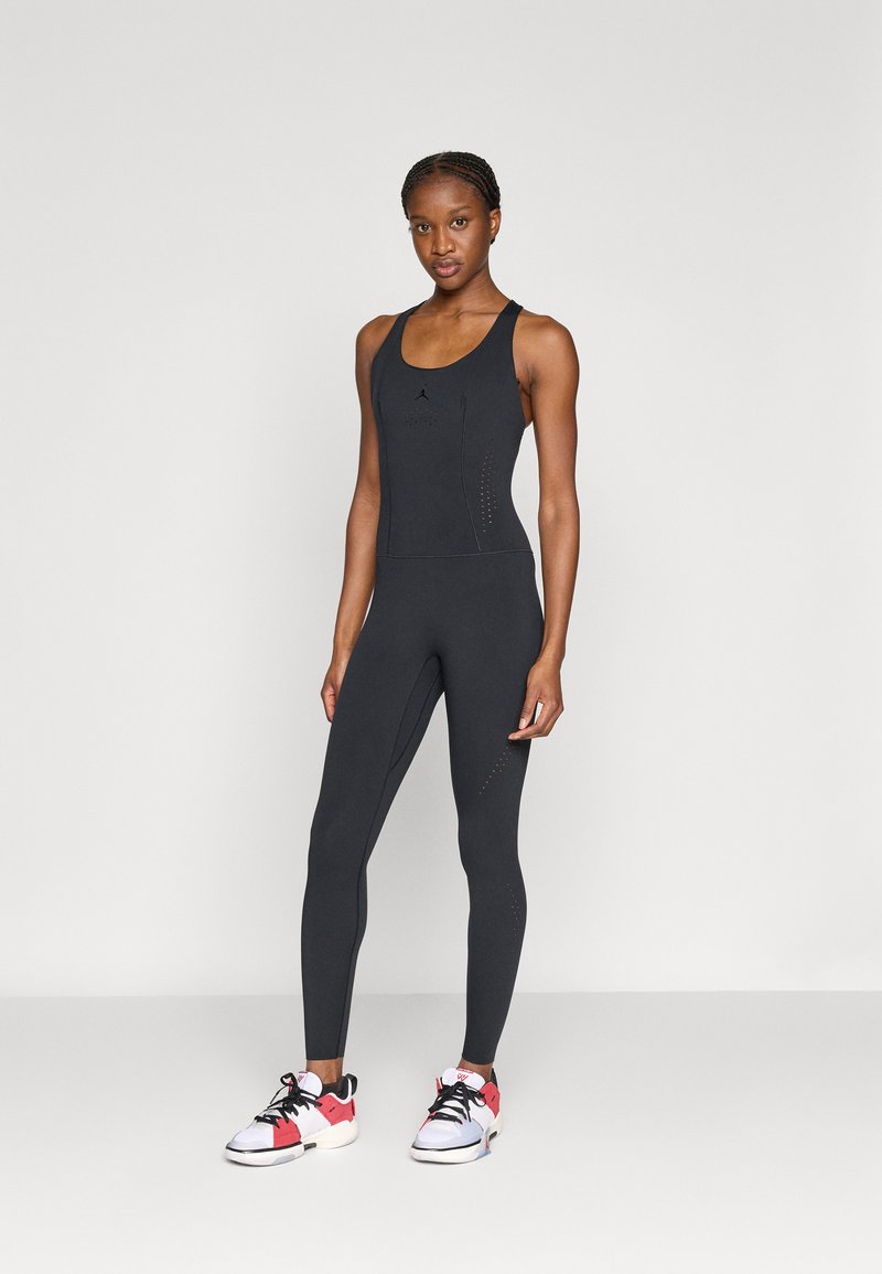 Justaucorps de sport noir avec un décolleté en U et un dos nageur. Présente des accents perforés et une coupe ajustée. Porté avec des chaussures de sport colorées.