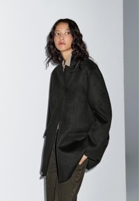 Czarny wełniany płaszcz o luźnym kroju, z oversize'owym kołnierzem, prostym krojem i dwoma przednimi kieszeniami. Na jasnoszarym tle.