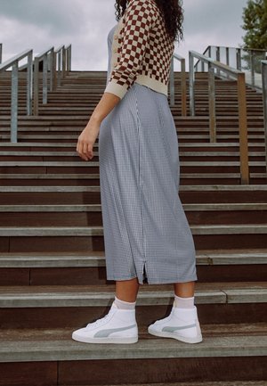 Personne debout sur des escaliers en bois extérieurs portant un cardigan à carreaux, une jupe à motifs noir et blanc et des baskets blanches.