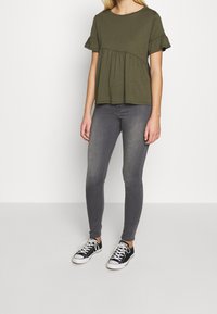 Olivově zelený krátký rukáv top s volánem, spárovaný se šedými skinny džíny a černými nízkými teniskami s bílou podrážkou.