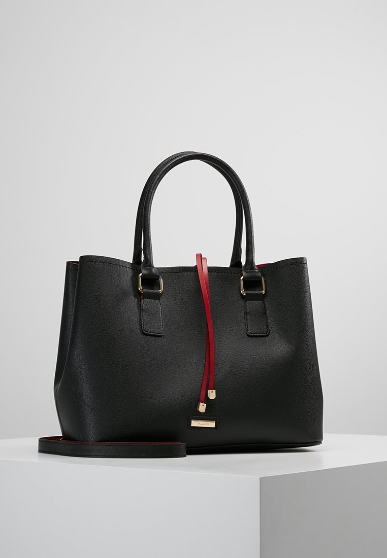 Sac à main en cuir noir avec double poignée, détail cordon rouge, accents dorés et bandoulière amovible posé sur une surface blanche.