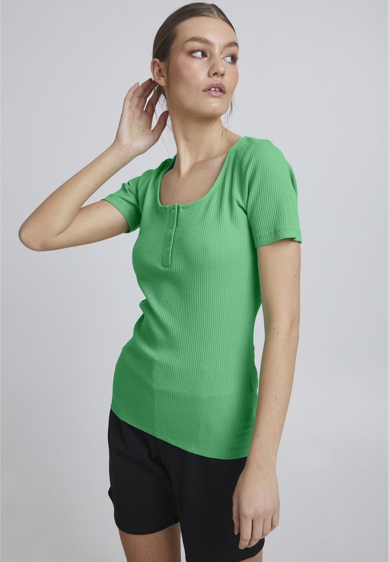 Top scurt cu maneci, de culoare verde, cu un decolteu rotund, închidere cu nasturi și un design ajustat, purtat cu pantaloni scurți negri.