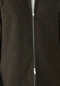 Hnedý vlnený kabát s vysokým golierom, so strieborným zipsom na prednej strane a mäkkým, štruktúrovaným povrchom. Žiadne viditeľné vrecká.