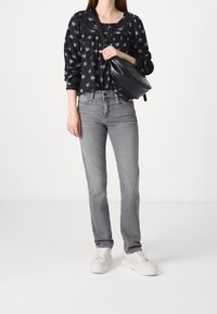 Femme portant un chemisier noir à motif floral, un jean gris droit, des baskets blanches, et tenant un sac bandoulière noir croisé sur sa poitrine.