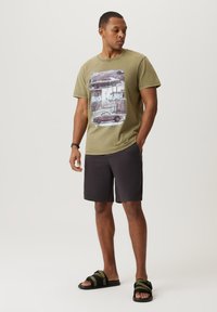 T-shirt w kolorze oliwkowym z nadrukiem vintage samochodu; zestawiony z ciemnymi szortami i czarnymi sandałami z zielonymi paskami. Swobodny, luźny krój.