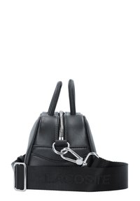 Borsa a mano in pelle nera con doppi manici superiori, chiusura con zip e tracolla in tessuto staccabile con il logo Lacoste. Texture liscia, forma minimalista.