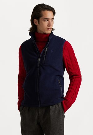 Navy fleece vest with high collar, featuring a zipper and side pockets, worn over a red knit turtleneck. Simple design, matte texture.