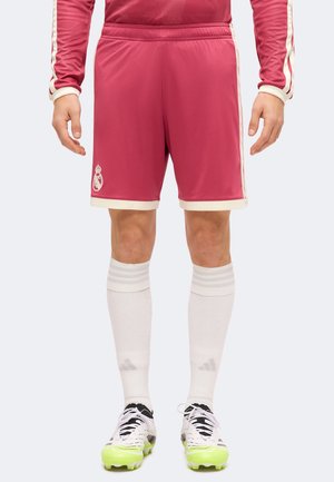 Hombre que lleva pantalones cortos de fútbol rosas con ribete blanco, calcetines blancos hasta la rodilla y botas blancas con suelas amarillo neón, de pie con los brazos a los lados.