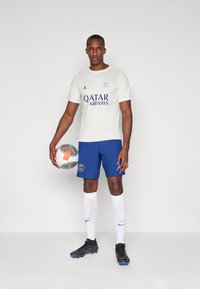 Fodboldudstyr bestående af en hvid trøje med blå tekst, blå shorts med strukturerede striber, hvide sokker og sorte støvler, som holder en mønstret bold.