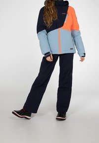 Farbblockierte Jacke mit navyblauen, hellblauen und orangefarbenen Abschnitten, mit einem Reißverschluss, kombiniert mit dunklen Hosen und schwarzen Schuhen.