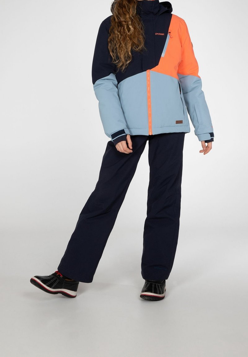 Farbblockierte Jacke mit navyblauen, hellblauen und orangefarbenen Abschnitten, mit einem Reißverschluss, kombiniert mit dunklen Hosen und schwarzen Schuhen.