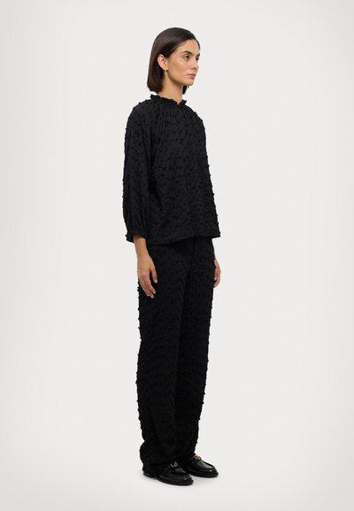 Sort tekstureret outfit med en løs bluse og vidde bukser, begge med et forhøjet mønster. Kombineret med sorte slip-on sko.