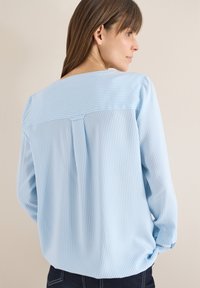 Femme de profil portant un chemisier à manches longues à rayures verticales bleu clair et blanc avec un pantalon foncé, sur un fond beige uni.