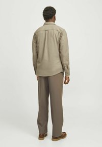 Chemise beige à manches longues avec poignets boutonnés, détail de l'empiècement au dos, associée à un pantalon marron ample et des chaussures marron.