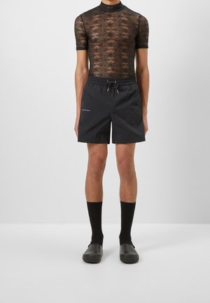 Personne portant un haut court en dentelle noire transparente à manches courtes, un short noir avec cordon de serrage, des chaussettes noires mi-mollet et des chaussures noires sans lacets, debout devant un fond blanc.