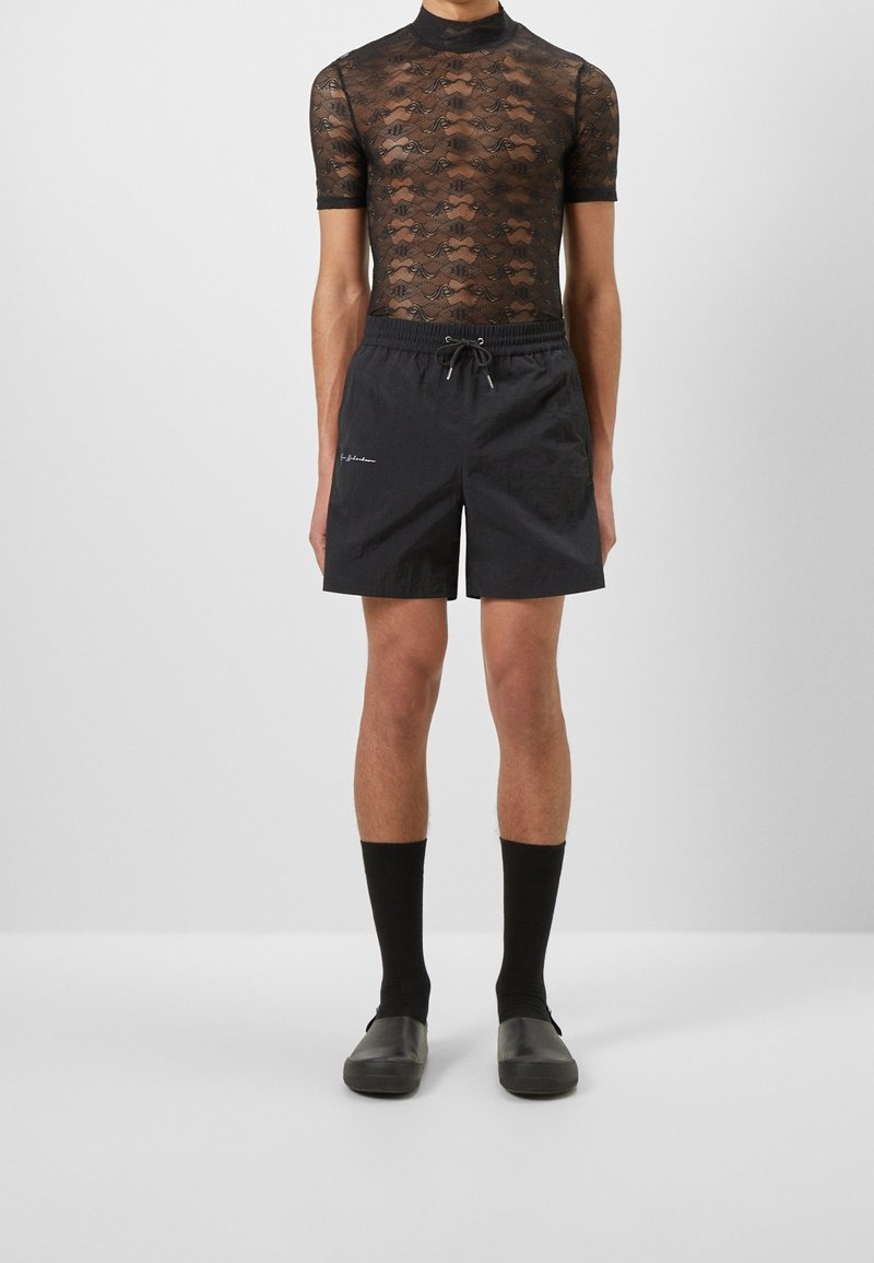 Personne portant un haut court en dentelle noire transparente à manches courtes, un short noir avec cordon de serrage, des chaussettes noires mi-mollet et des chaussures noires sans lacets, debout devant un fond blanc.