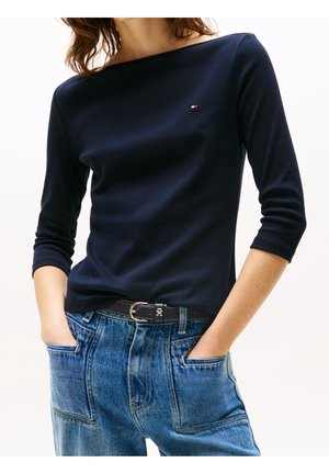 Femme portant un haut à manches longues bleu marine avec un petit logo, rentré dans un jean bleu taille haute avec une ceinture noire, mains dans les poches.