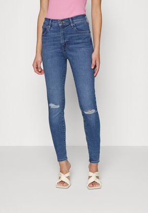 Femme portant un jean skinny bleu taille haute avec des genoux déchirés, un top rose sans manches et des sandales blanches à talons avec des brides croisées.