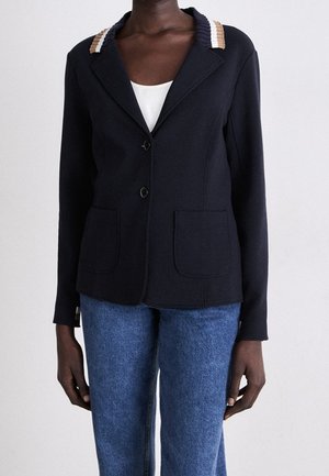 Granatowa marynarka z dwoma przednimi kieszeniami, dwoma guzikami oraz beżowo-paskowym zdobieniem na ramionach, noszona na biały top i niebieskie dżinsy.