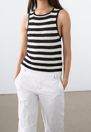 Vrouw met een mouwloos, zwart-wit horizontaal gestreept gebreid top gecombineerd met losse witte broek, handen in de zakken, binnen.