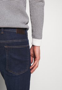 Mörkblå denimjeans med rak passform, som har en brun läderlapp på baksidan av midjan och kontrasterande sömmar på fickorna.
