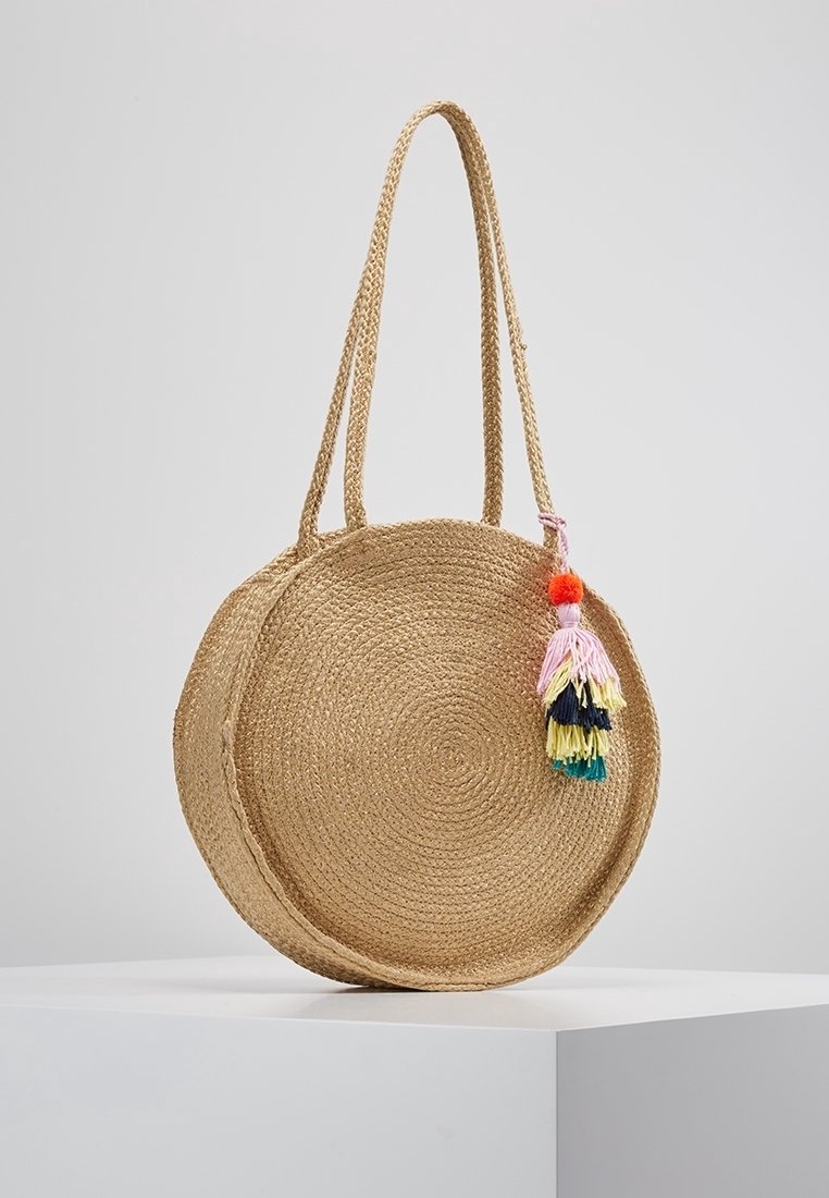 Sac à main rond en paille tressée avec de longues anses et une décoration de pompons colorés suspendue d'un côté.