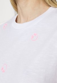 Primer plano de una camiseta blanca con pequeños diseños de caras sonrientes rosas impresos cerca del cuello y la zona del hombro.