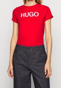 Červené bavlněné tričko s ohrnutými rukávy, na přední straně s velkým bílým logem "HUGO". Spárované s tmavými džíny, zdůrazňujícími kontrasty.