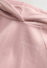 Růžová mikina s kapucí vyrobená z měkké bavlny, s přední kapsou a jemným logem v bílé barvě. Obsahuje šňůrku na stažení a žebrované manžety.