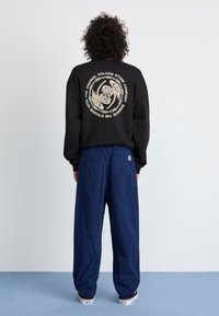 Sudadera negra con diseño gráfico beige en la parte trasera, combinada con pantalones sueltos azul marino y zapatos negros. Fondo liso.