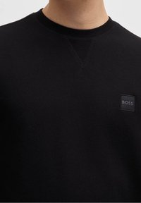 Sudadera negra confeccionada con tela suave que presenta un cuello redondo, puños acanalados y un pequeño parche con logo cuadrado en el pecho.