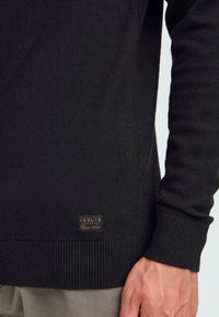 Pulover negru din tricot, cu tiv și manșete cu dungi. Include un mic eticheta cu logo texturat în partea stângă jos. Textură netedă pe toată suprafața.