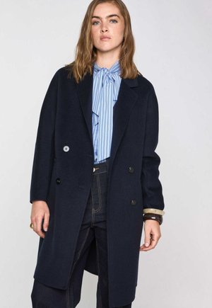Marineblauwe wollen blend dubbelzijdige jas met grote knopen, notch revers en zijzakken, over een lichtblauw gestreept blouse en donkere jeans.