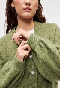 Πράσινη πλεκτή ζακέτα με υφή, με γιακά σάλι και λεία, ανοιχτόχρωμα κουμπιά. Το μοντέλο ποζάρει με τα χέρια κοντά στα μανίκια.